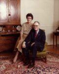 a woman sits on the arm of a chair while the man sits in the chair both are smiling at the camera