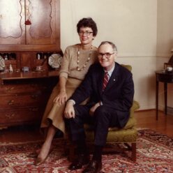 a woman sits on the arm of a chair while the man sits in the chair both are smiling at the camera