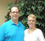 a man wearing a blue shirt and a woman wearing a white shirt stand in front of a faux tree smiling at the camera