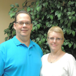 a man wearing a blue shirt and a woman wearing a white shirt stand in front of a faux tree smiling at the camera