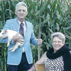 Smiling man holding a piglet, and smiling woman holding a hat on her lap