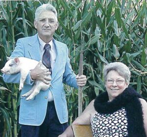 Smiling man holding a piglet, and smiling woman holding a hat on her lap