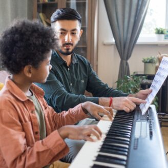 A man holds a sheet of music as he sits with a child at a keyboard.