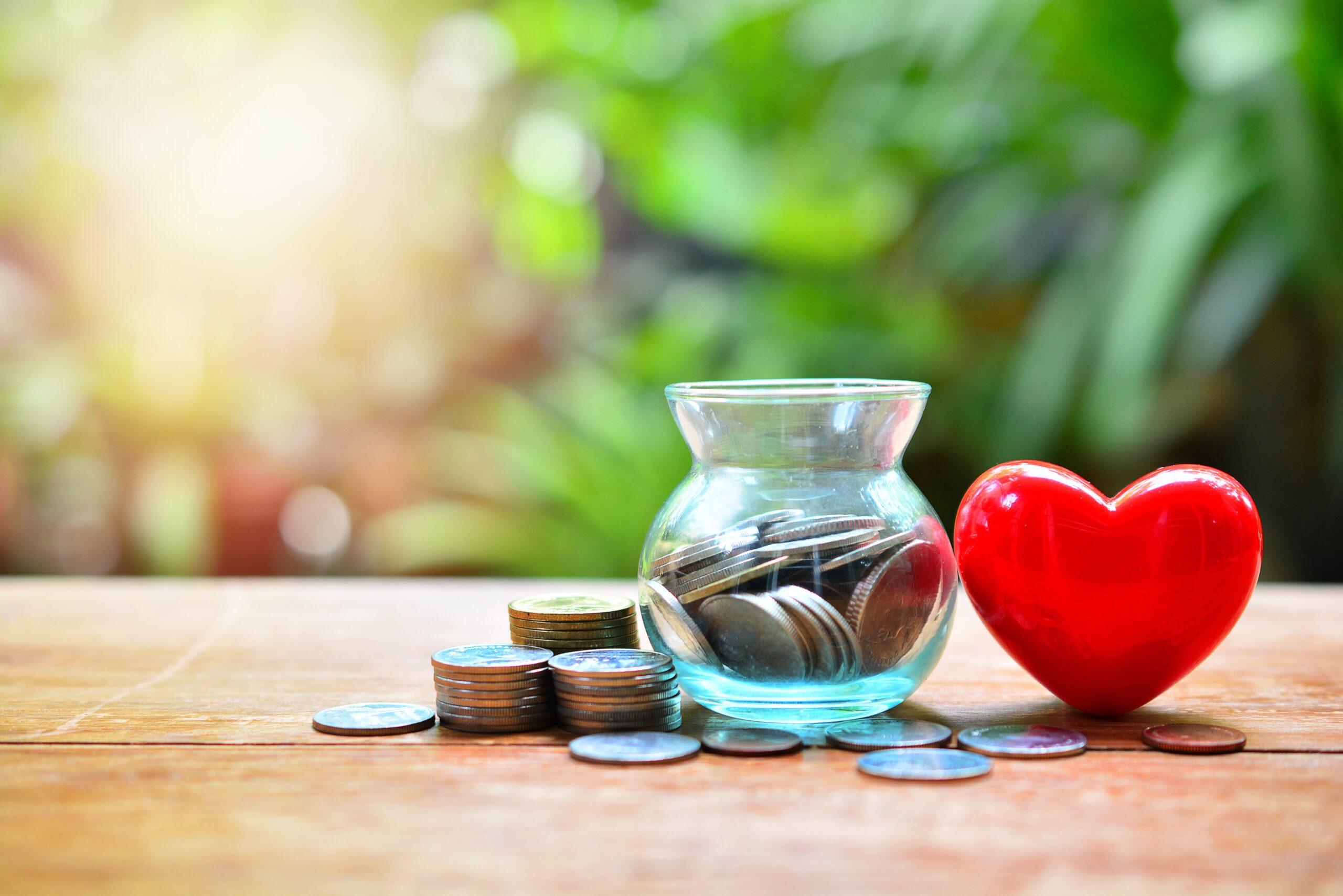 A jar with coins in it sits on a table next to a red heart and other loose coins.