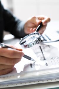 A close up of hands holding a pen and magnifying glass looking at a financial document. 