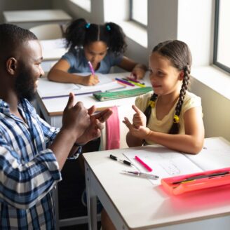 A male teacher kneels down to speak in sign language to a young student.