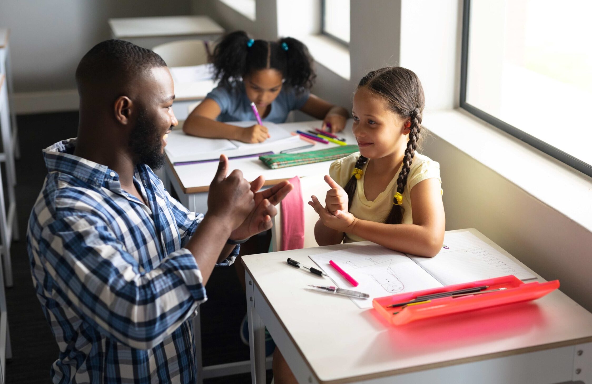 A male teacher kneels down to speak in sign language to a young student.