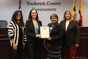 Four professional women pose for a photo while holding a proclamation in a county office building. 