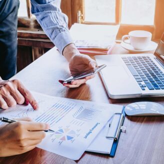 Two people sit at a desk holding a phone, laptop and financial documents.