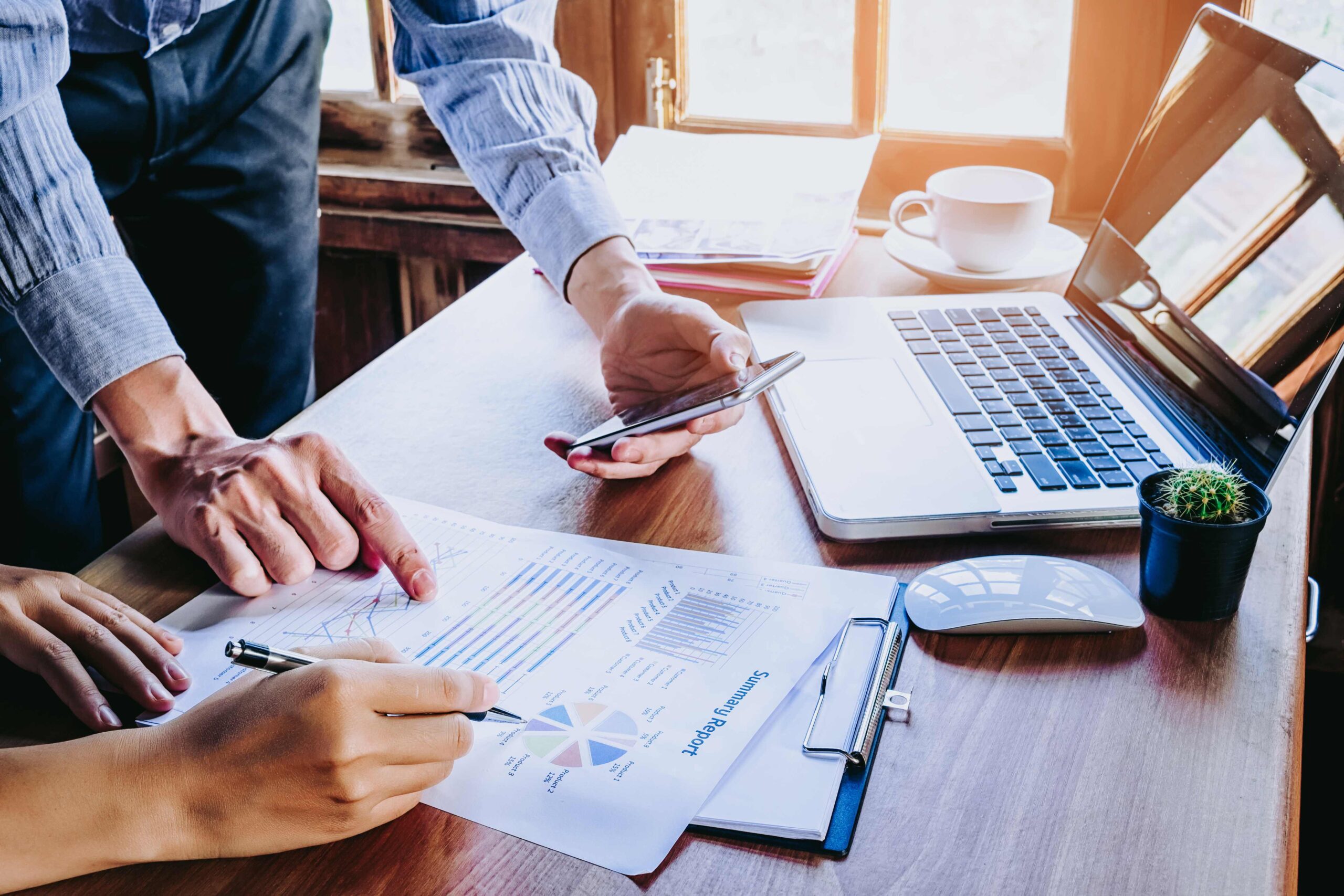 Two people sit at a desk holding a phone, laptop and financial documents. 