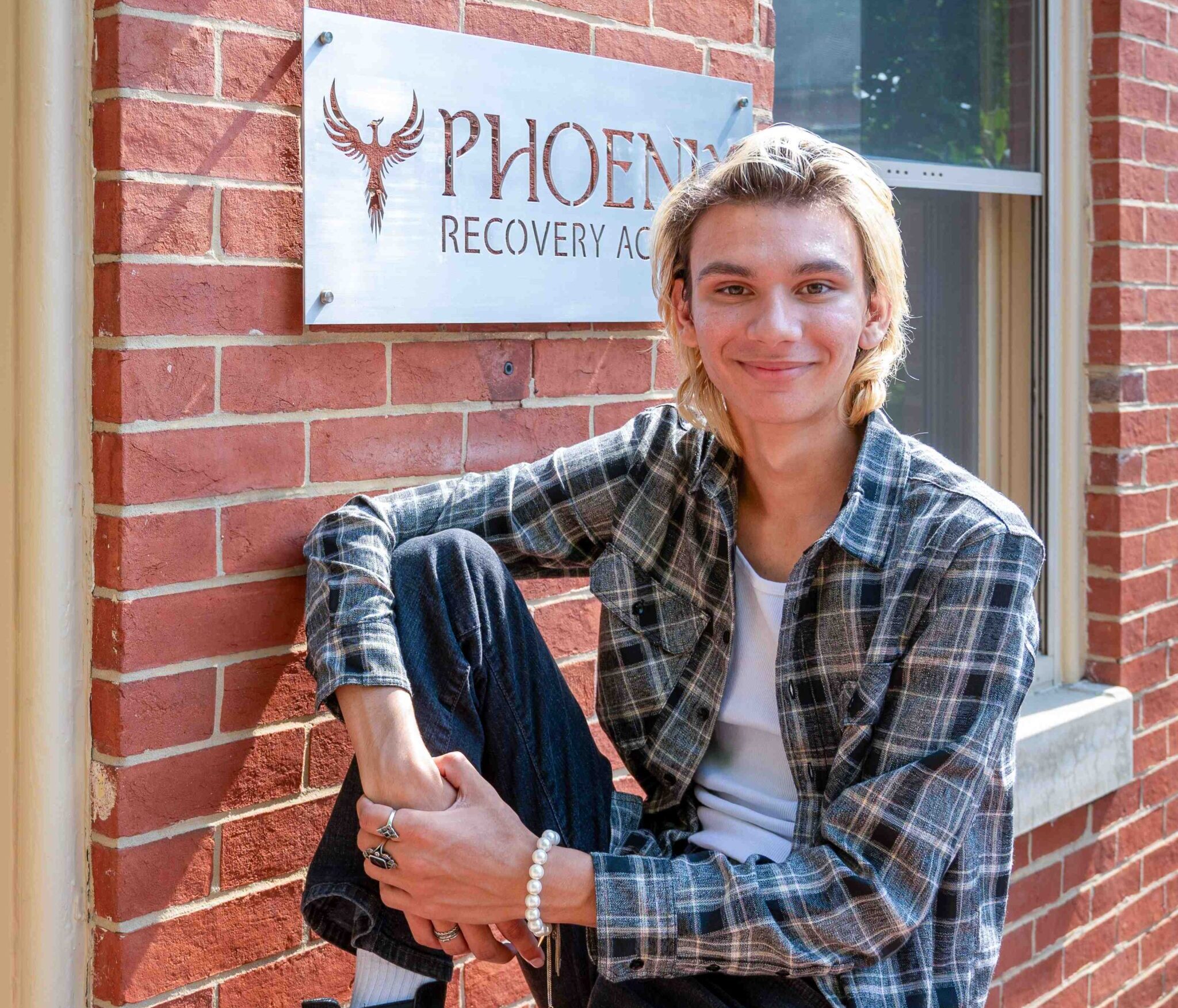 A young man sits on a brick porch in front of a sign that reads "Phoenix Recovery Academy". 