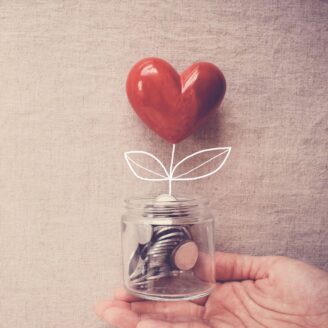 A hand holds a jar with coins in it and a heart growing out of the top.