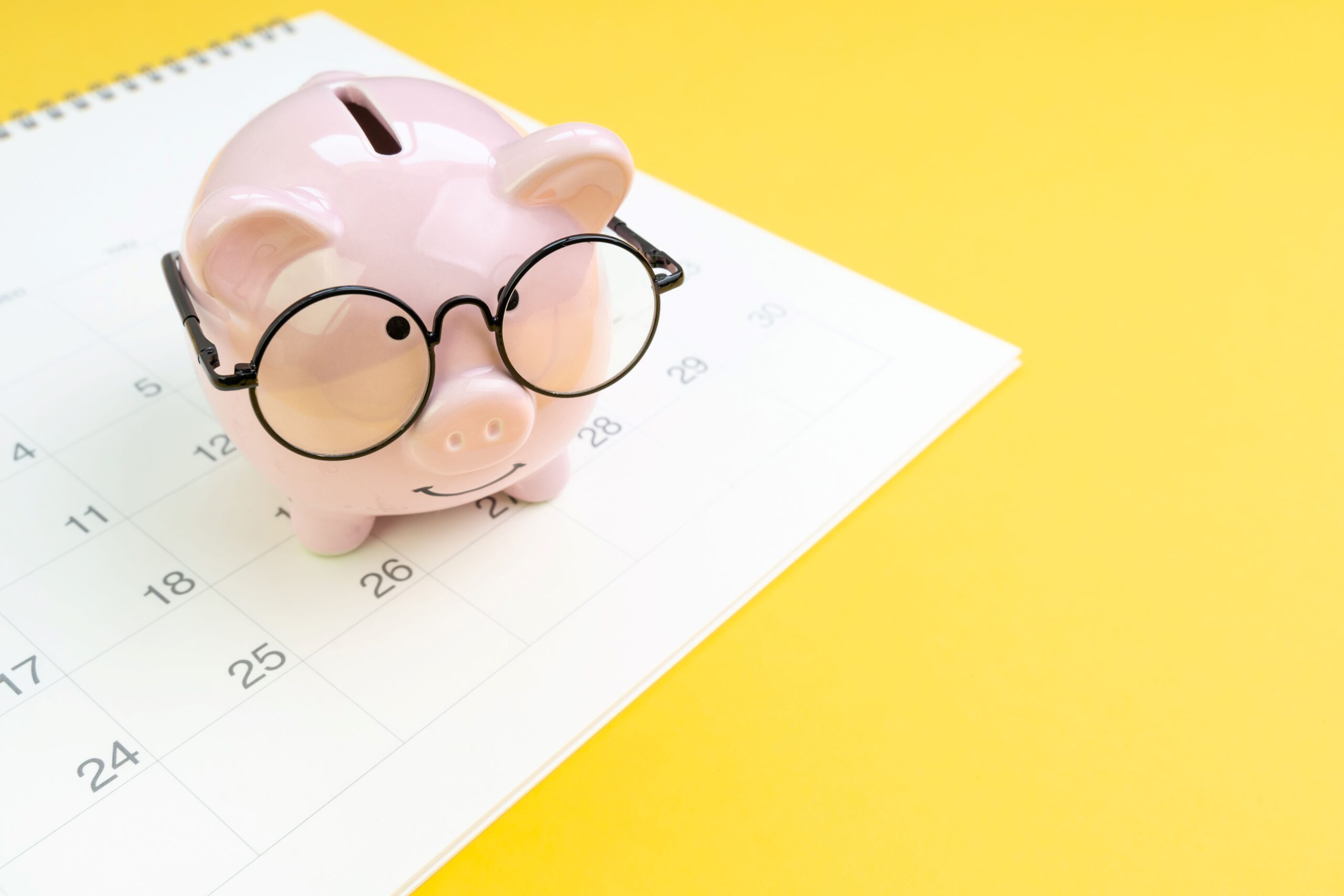 A piggy bank with glasses on sits on top of a calendar. 