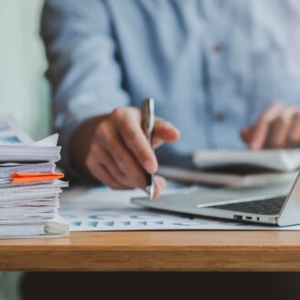 person at a desk working on a laptop with a stack of papers next to them