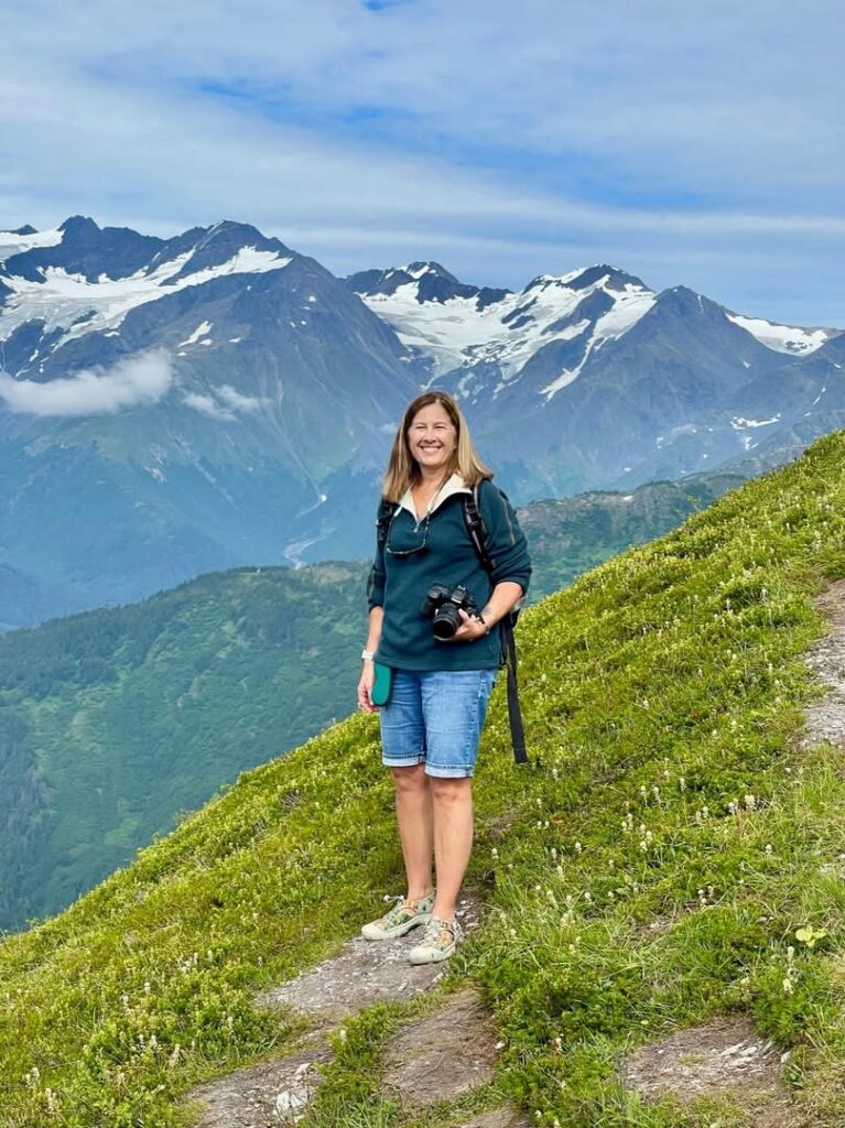 Woman stands on a trail with mountain range behind her
