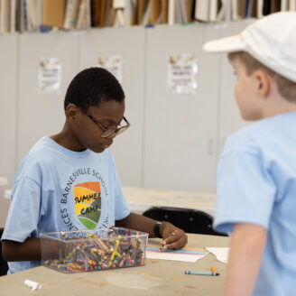 child sits coloring at a table with a large container of crayons in front of him